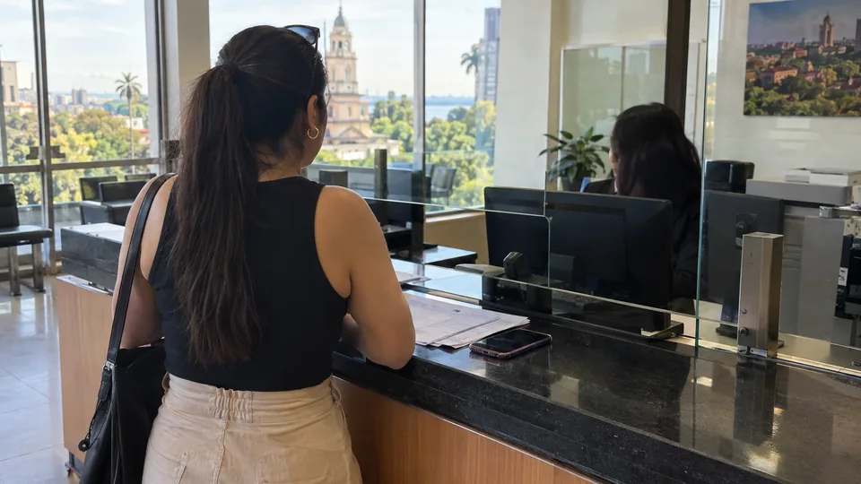 Mulher de costas no balcão de atendimento de uma agência bancária em Assunção, com papéis e celular sobre o balcão, em imagem realista gerada por IA