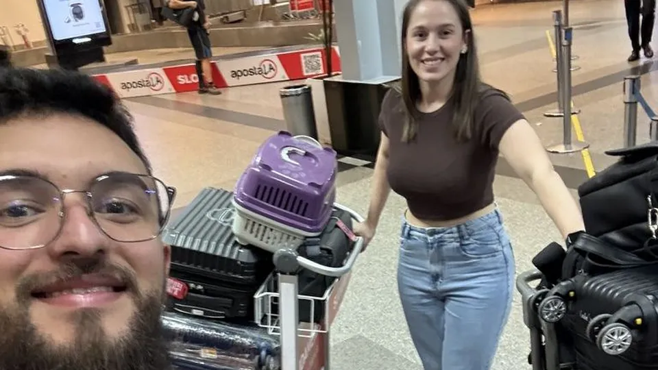 Samuel sorrindo com sinal de positivo no aeroporto, com Pâmela e as malas ao fundo