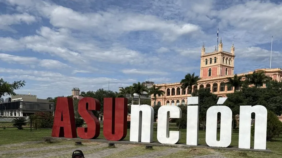 Samuel agachado em frente ao letreiro colorido de Assunção
