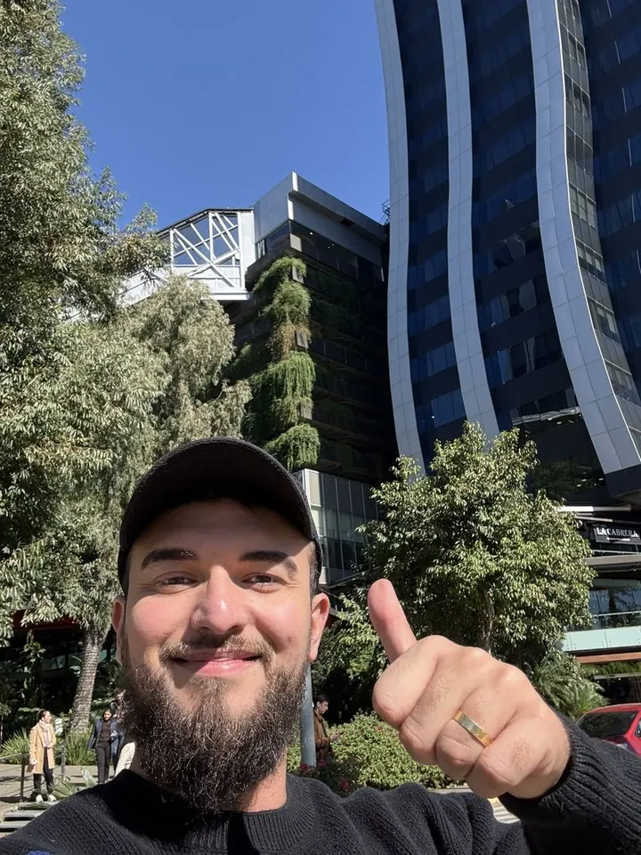 Samuel fazendo sinal de positivo em frente a prédios espelhados e árvores