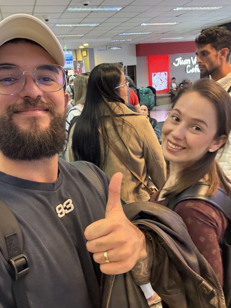 Samuel e Pâmela em uma selfie sorridente, ele fazendo sinal de positivo, em um prédio público cheio de gente