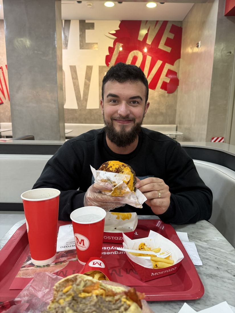 Ele sorrindo com um lanche na mão em uma lanchonete